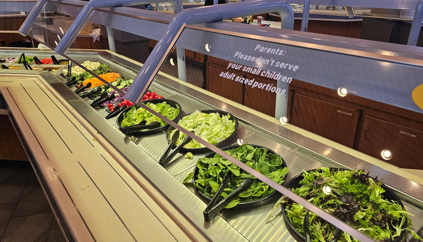 A salad bar with various greens, vegetables, and toppings in black bowls is featured at the all-you-can-eat breakfast buffet. A sign reads, "Parents: Please don't serve your small children adult sized portions." Brown cabinets sit in the well-lit background.