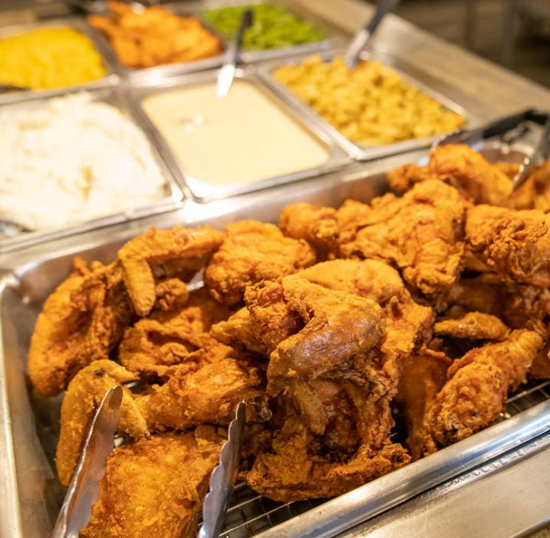 A tray of crispy fried chicken sits in front of an all-you-can-eat breakfast buffet with mashed potatoes, gravy, macaroni and cheese, green beans, and other sides. Metal tongs rest on the tray for serving.