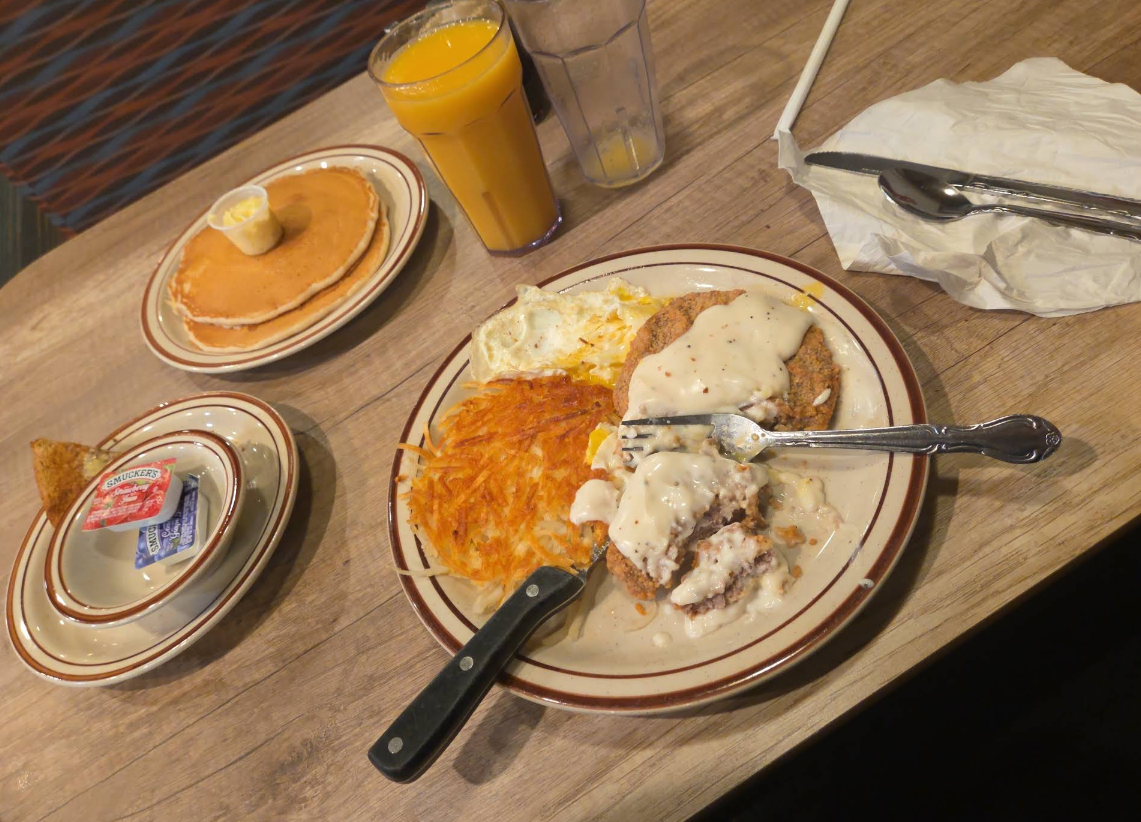A classic diner all-you-can-eat breakfast buffet featuring pancakes, orange juice, hash browns, eggs, and chicken-fried steak covered in white gravy. Toast with jelly, utensils, and a napkin complete the feast.