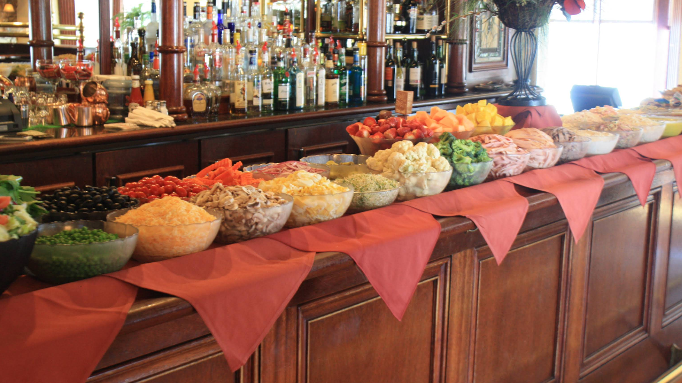 A long bar counter displays bowls of assorted salad ingredients, including vegetables, meats, cheeses, and fruits, arranged in rows with red napkins draped along the edge—perfect for an all-you-can-eat breakfast buffet. Bottles and glasses fill the shelves behind.