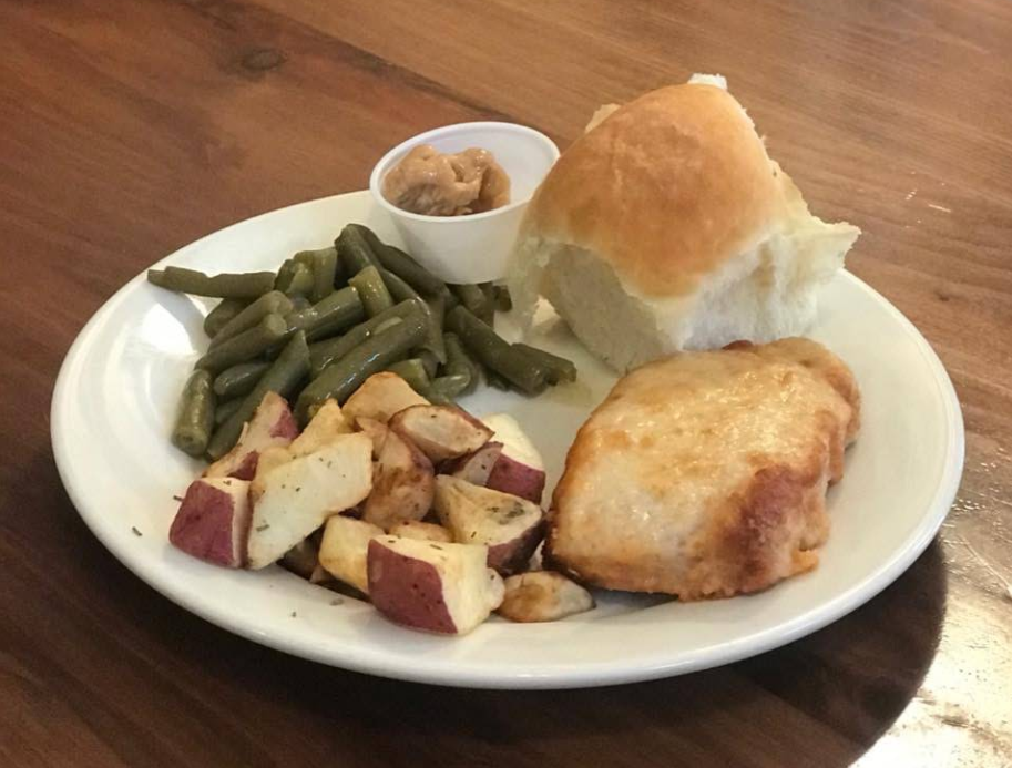 A dinner plate with baked chicken breast, roasted red potatoes, green beans, a dinner roll, and a small cup of apple butter on a wooden table—perfect after enjoying our all-you-can-eat breakfast buffet.
