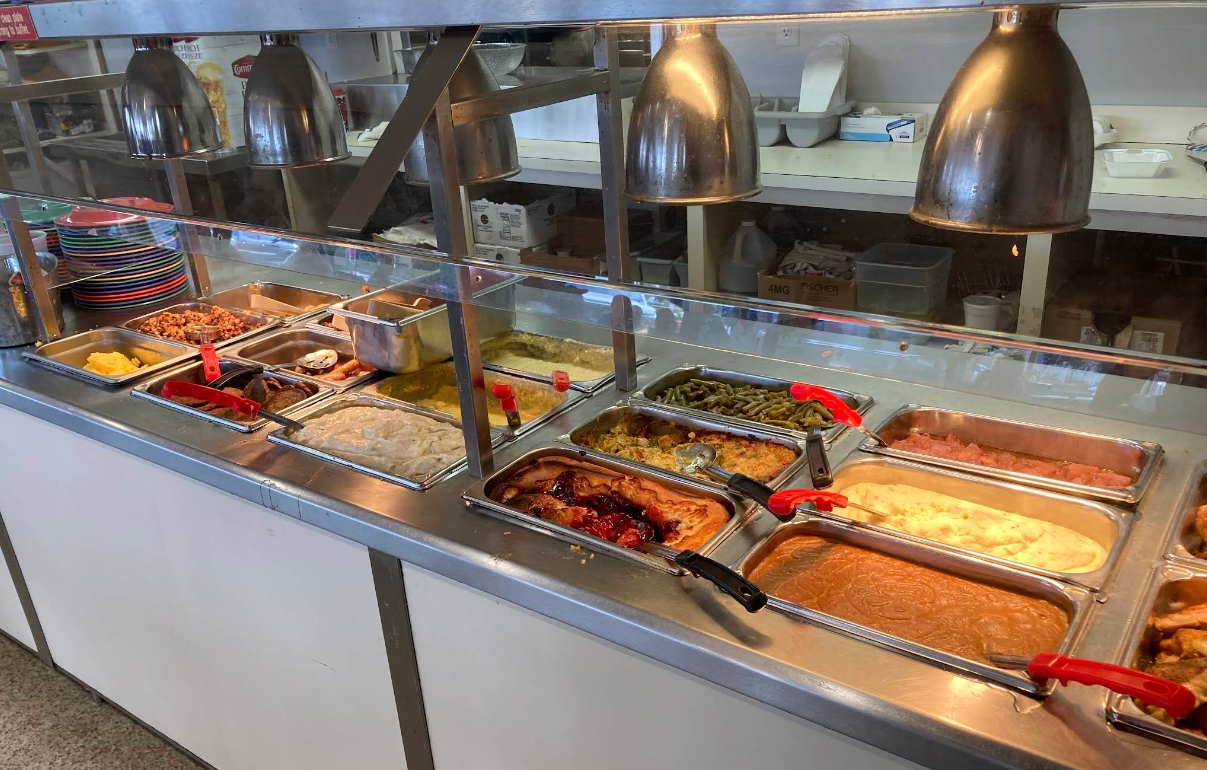 A cafeteria buffet features a variety of hot foods, including green beans, mashed potatoes, corn, fried chicken, and other sides—all part of the all-you-can-eat breakfast buffet displayed in stainless steel trays under heat lamps.