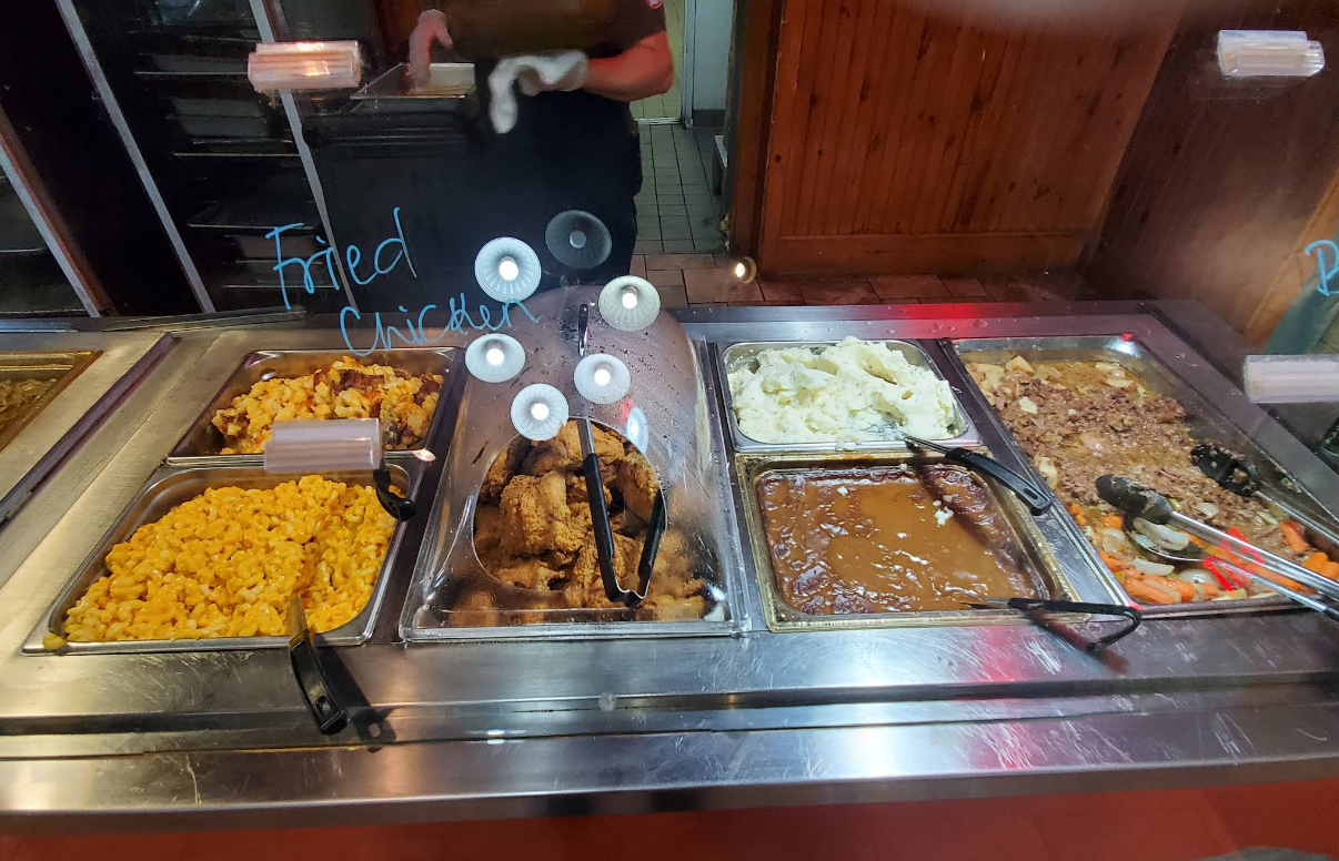 A cafeteria-style, all-you-can-eat breakfast buffet with trays of macaroni and cheese, fried chicken, mashed potatoes with gravy, and another meat dish behind a glass divider. A person’s hand is visible behind the counter.