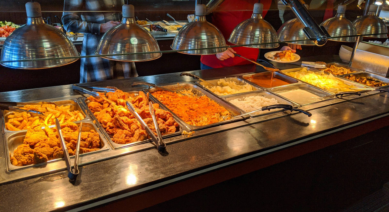 An all-you-can-eat breakfast buffet features trays of fried chicken, French fries, carrots, mashed potatoes, rice, macaroni and cheese, and other hot foods. Two people are serving themselves under warm lights.