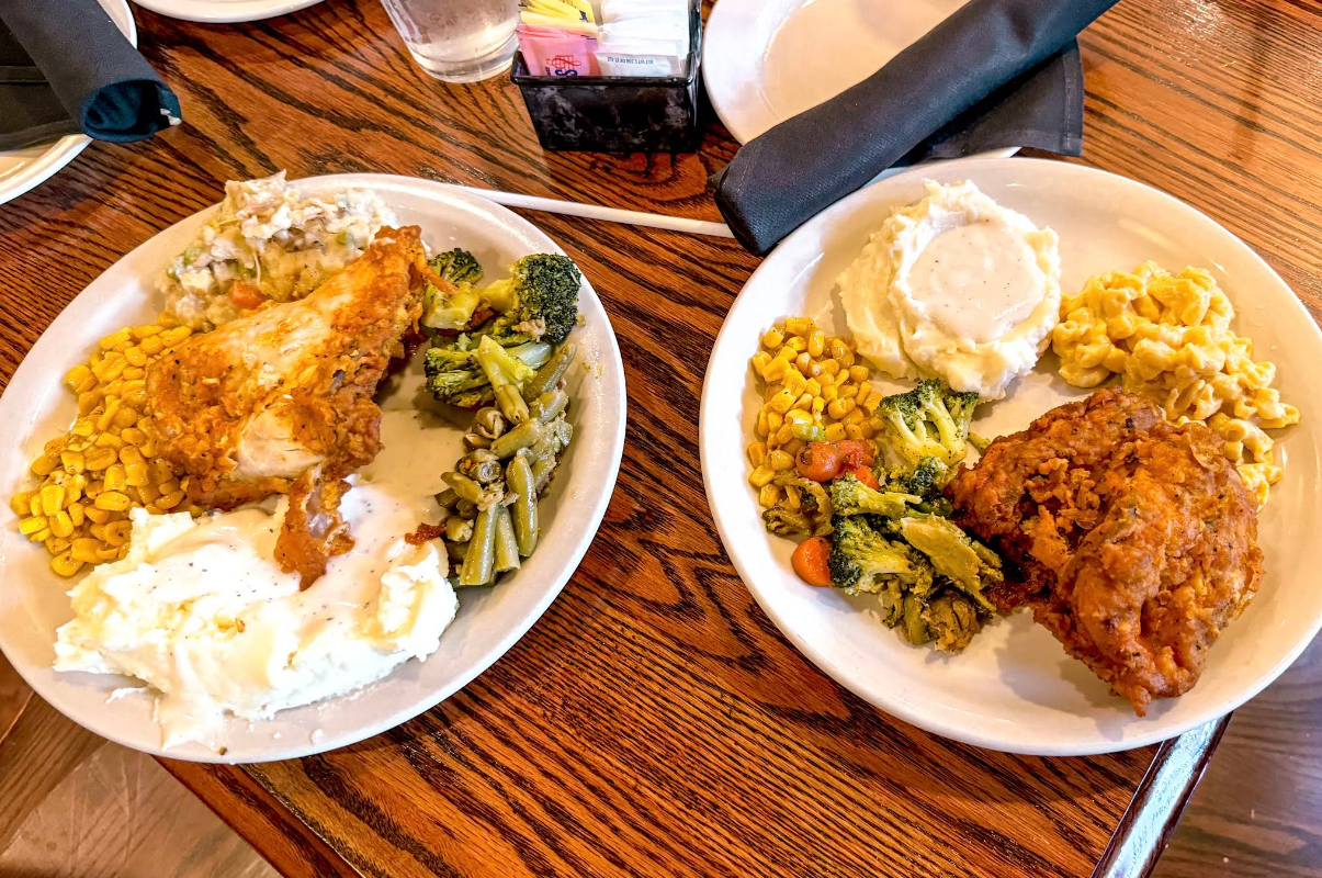 Two plates of food on a wooden table, each with fried chicken, mashed potatoes with gravy, corn, broccoli with carrots, and one plate also features macaroni and cheese—like a hearty all-you-can-eat breakfast buffet. Black napkin and drink cup in the background.