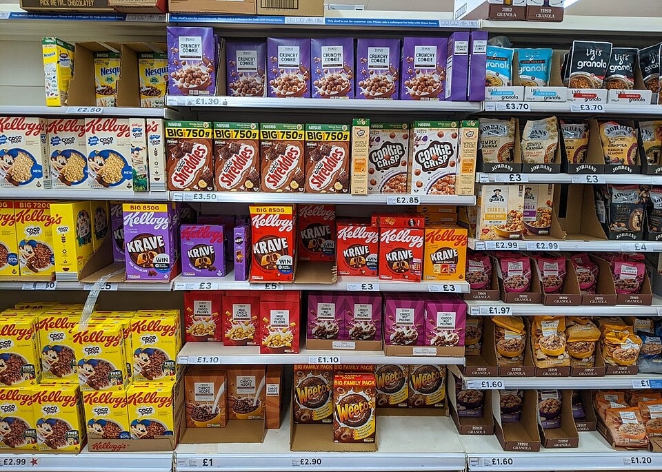 Shelves in a supermarket display various boxed cereals, including Cookie Crisp, Krave, Weetos, and Nesquik, with colorful packaging and price labels visible below each product.