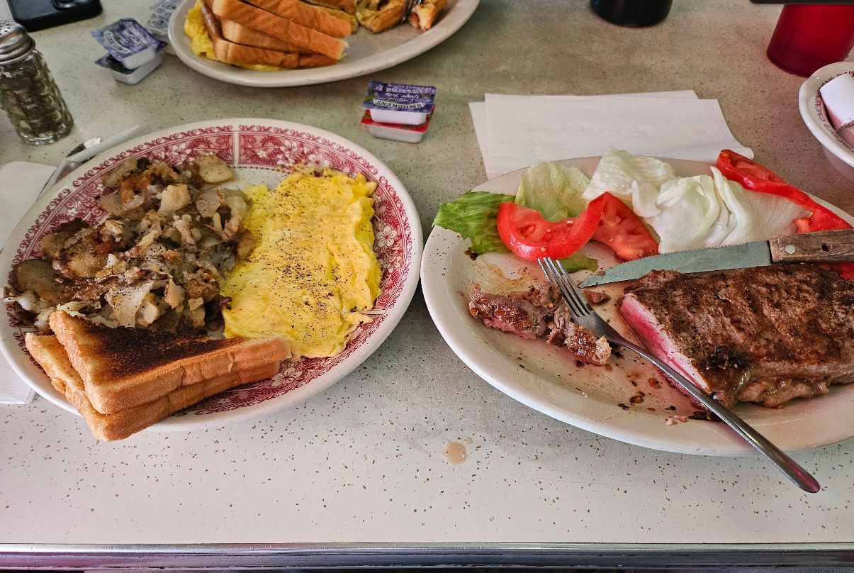 Two plates from an all-you-can-eat breakfast buffet: one with scrambled eggs, home fries, and toast; the other with a partially eaten steak, tomato, lettuce, and a steak knife. Butter packets and napkins are also visible.