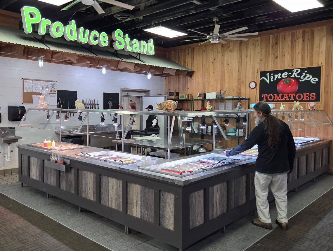 A person stands in front of a self-serve counter at an indoor produce stand, which is empty except for a few items. Signs above read "Produce Stand" and "Vine-Ripe Tomatoes," evoking a rustic charm—unlike an all-you-can-eat breakfast buffet.