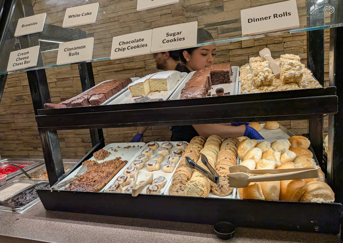 A bakery display case with brownies, cinnamon rolls, white cake, chocolate chip cookies, sugar cookies, cream cheese bars, and dinner rolls. A woman is behind the counter serving customers at the all-you-can-eat breakfast buffet.