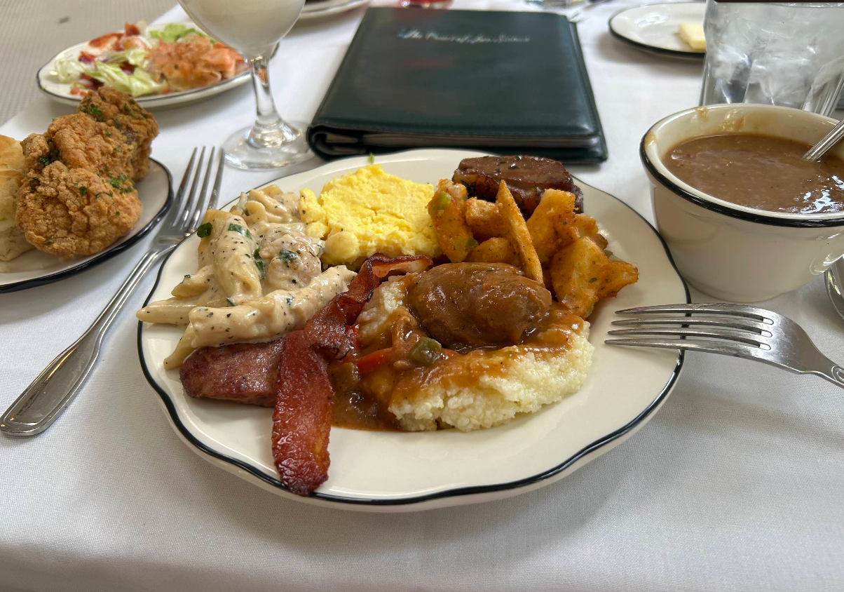 A plate from the all-you-can-eat breakfast buffet features bacon, scrambled eggs, creamy chicken, roasted potatoes, sausage, meat over grits with gravy. A bowl of soup, fried chicken, and salad are nearby on a white tablecloth.