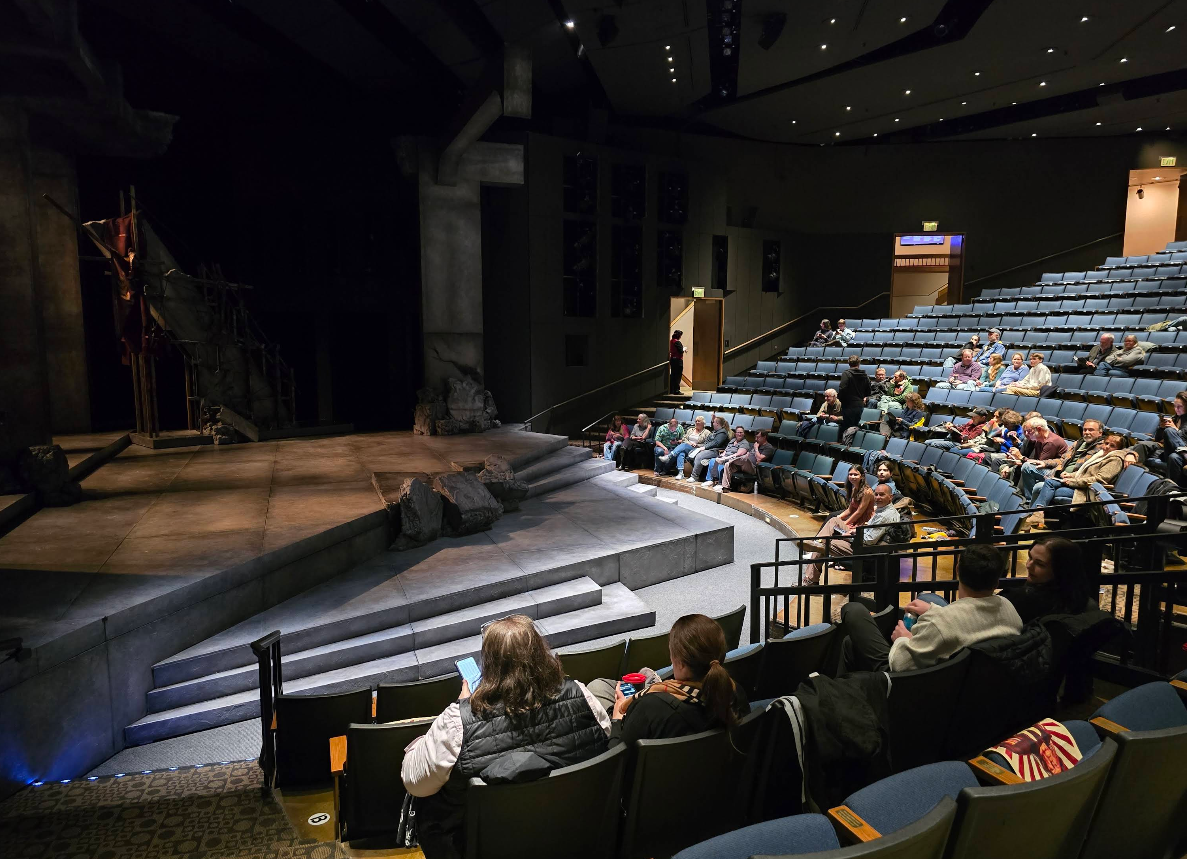 Audience members sit in a live theater with blue seats, facing a stage set with rocky scenery. The house lights are on, the theater is partially filled, and the stage is currently empty.