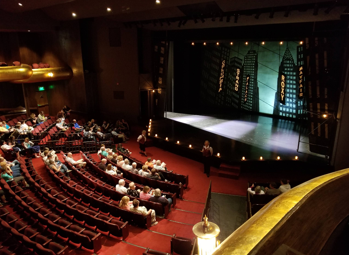 A live theater with red seats and an audience waiting for a show to begin. The stage is set with tall, cityscape cutouts and spotlights, as two people stand near the edge of the stage addressing the crowd.