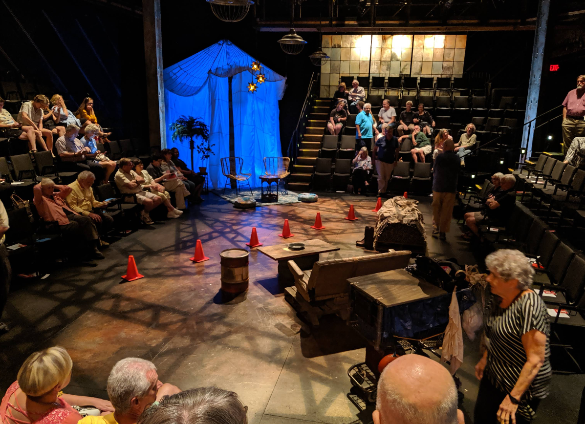 Audience members are seated in a dimly lit live theater, with a set resembling an outdoor scene, featuring orange cones, props, and a blue-lit tent at center stage. People chat and wait for the performance to begin.