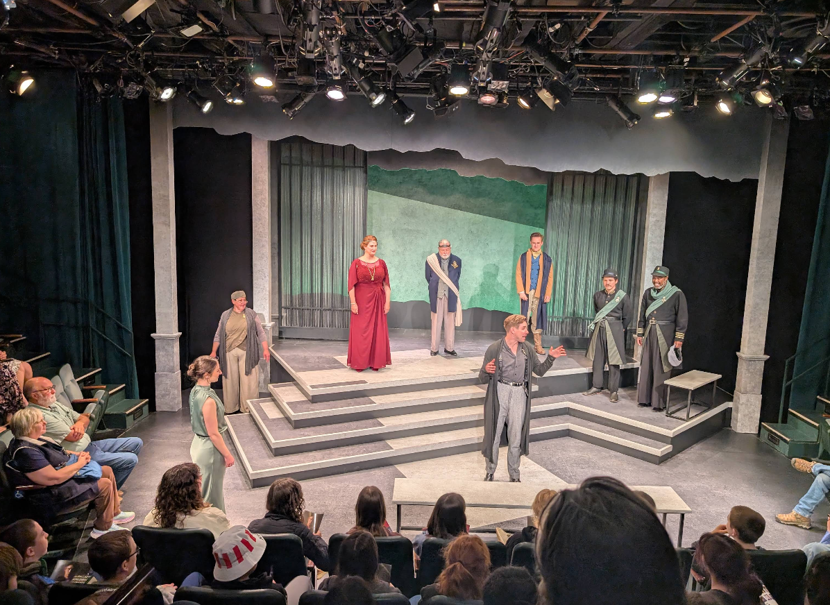 Actors in period costumes perform a dramatic scene on a tiered stage at this live theater event, with an audience watching. The set features columns, steps, and a painted backdrop, while lighting highlights the performers.