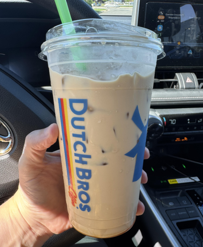A hand holds a large iced coffee from one of the popular coffee houses, Dutch Bros, in a clear cup with a green straw inside a car; the dashboard, steering wheel, and digital display are visible in the background.