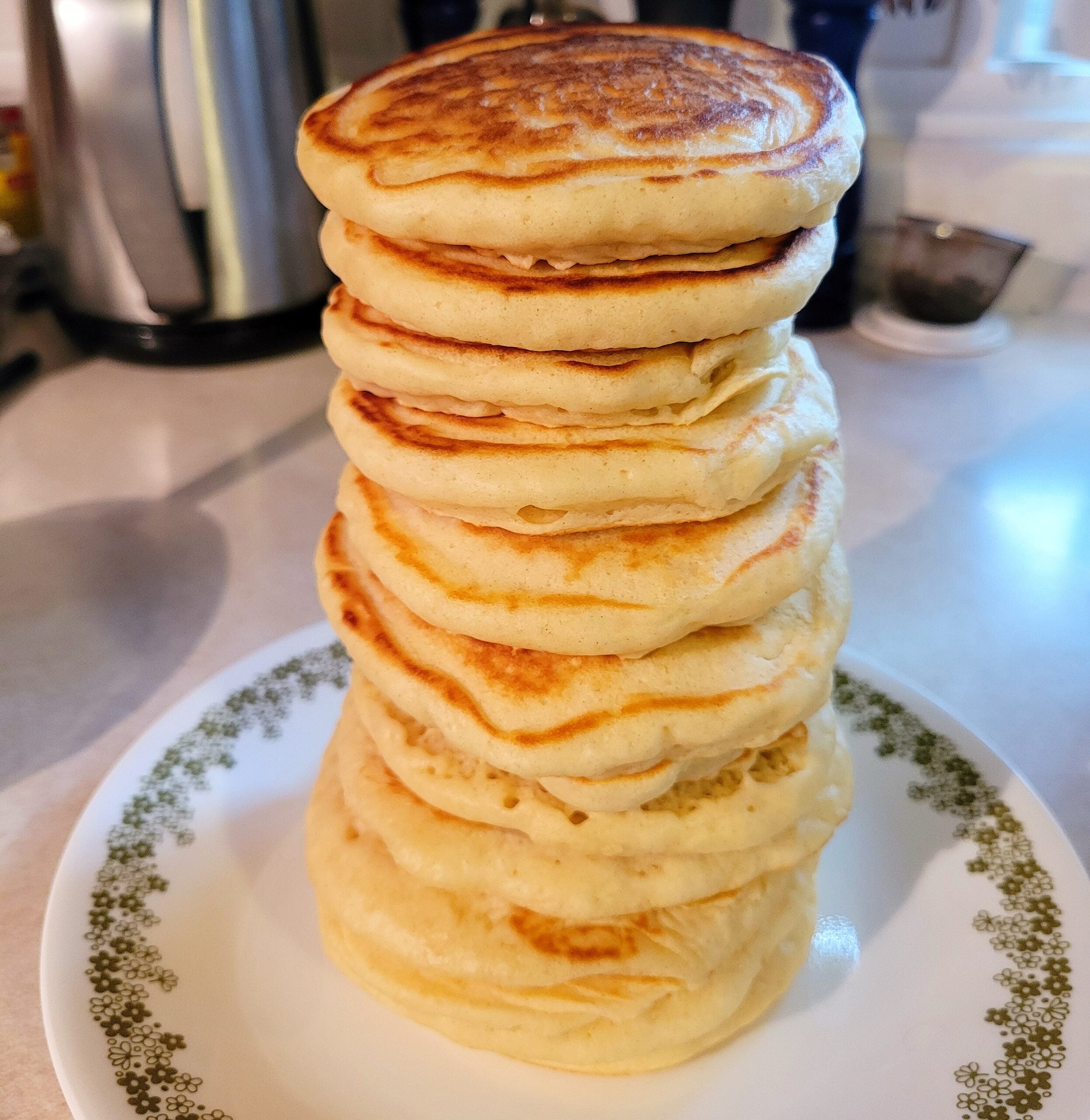 A tall stack of golden, fluffy homemade pancakes is neatly arranged on a white plate with a green floral border, sitting on a kitchen counter.