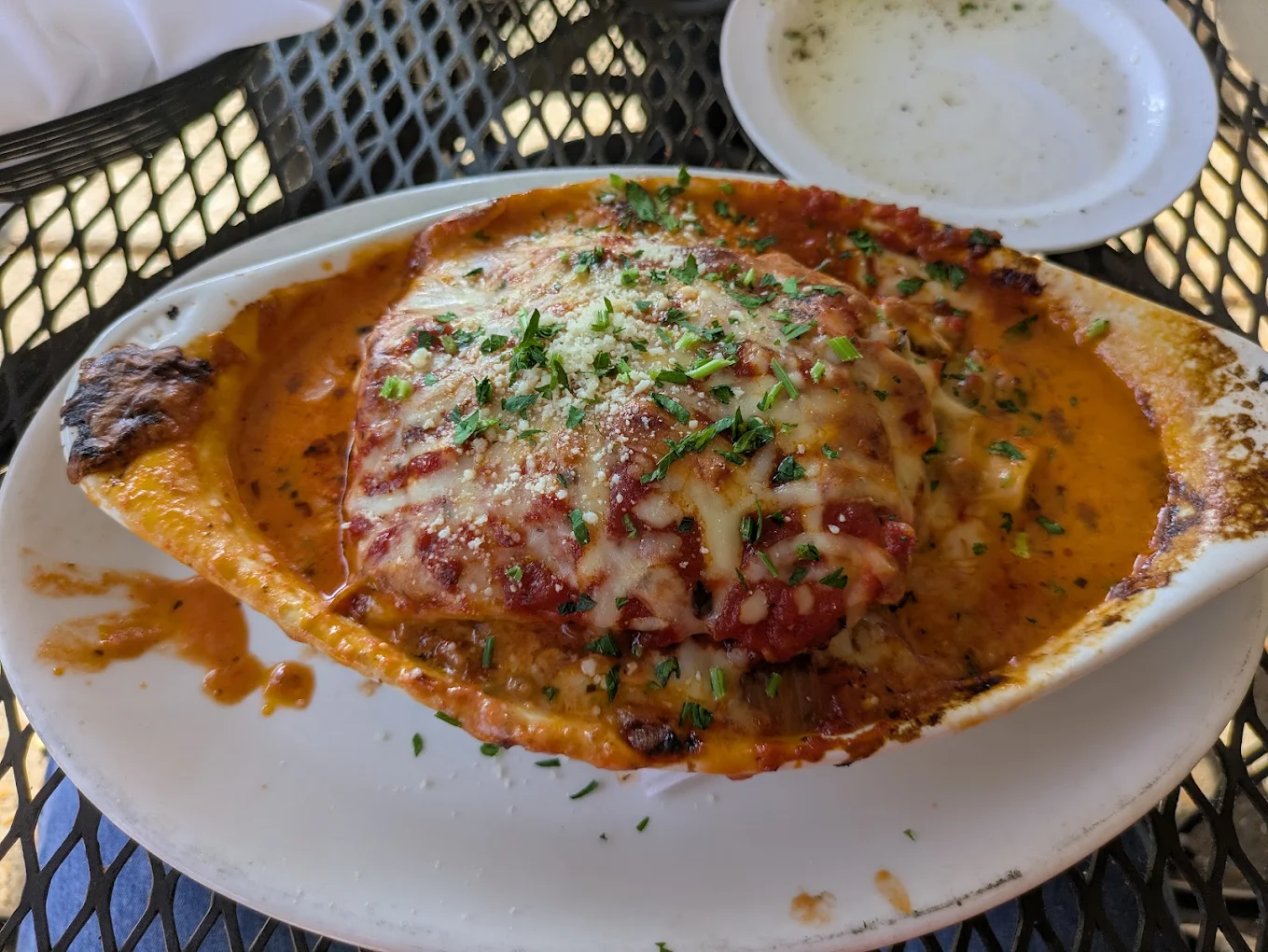 Baked lasagna topped with melted cheese, tomato sauce, and herbs served in an oval dish on a white plate, with a small bowl of olive oil and herbs in the background.