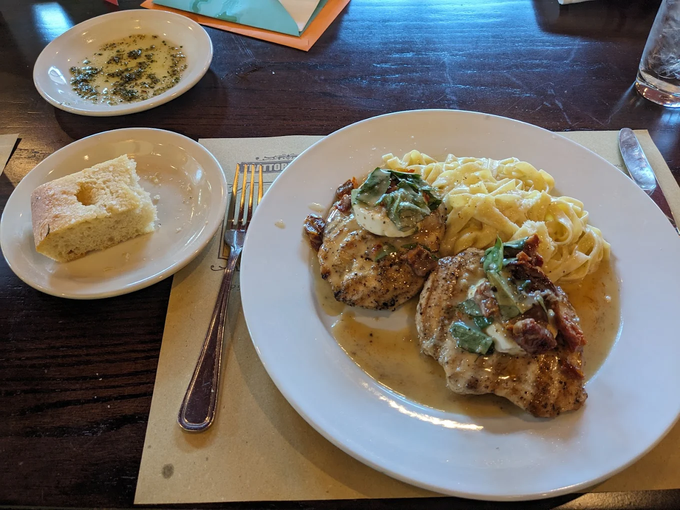 A plate with pasta, two pieces of grilled chicken topped with sauce and herbs, and a side dish of cornbread with a fork and knife. A small bowl with a dip or dressing is in the background.