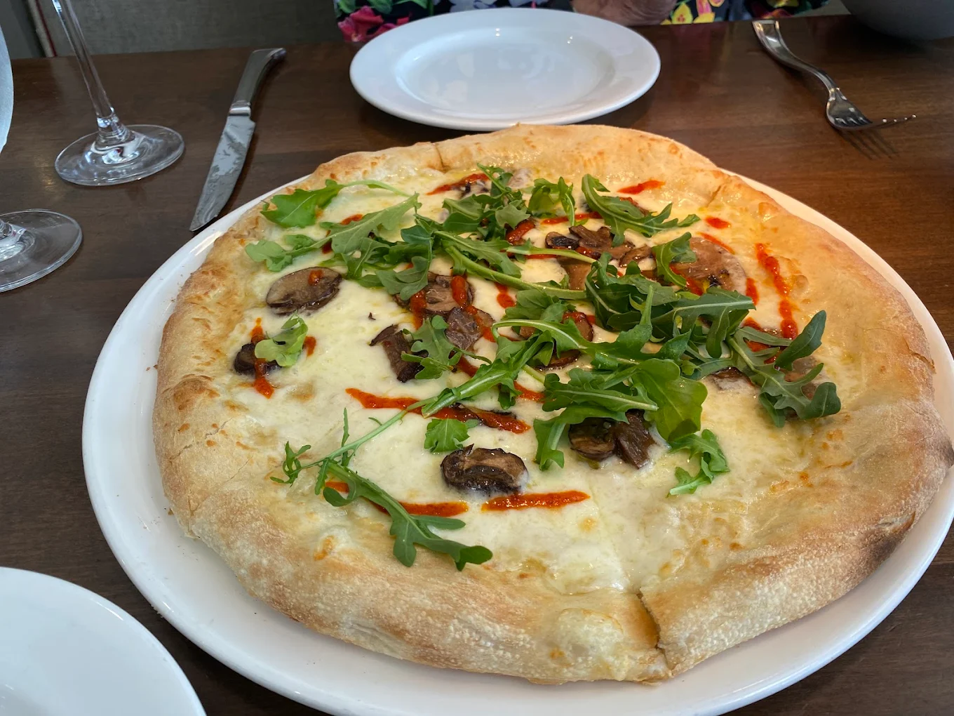 A pizza topped with arugula, mushrooms, cheese, and a drizzle of red sauce sits on a white plate on a wooden table, with an empty plate, utensils, and a glass nearby.