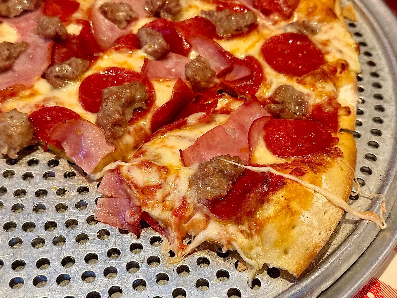 A close-up of a meat lover’s pizza with pepperoni, sausage, and ham on a perforated metal tray. One slice is partially pulled away, stretching melted cheese.