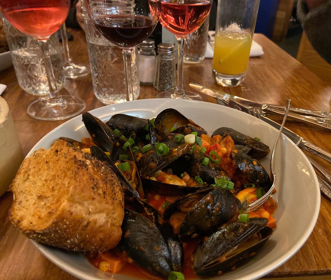A bowl of mussels in a tomato-based sauce garnished with green onions, served with a slice of toasted bread. Glasses of red and rosé wine, water, and orange juice are on the wooden table.