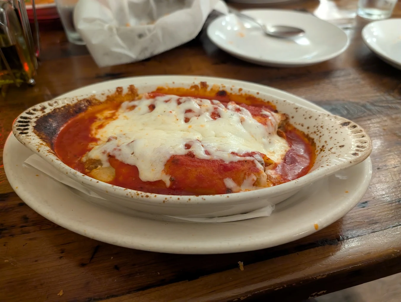 A baked dish of lasagna topped with melted cheese and tomato sauce sits on a white plate on a wooden table, with empty plates, a spoon, and a bread basket in the background.