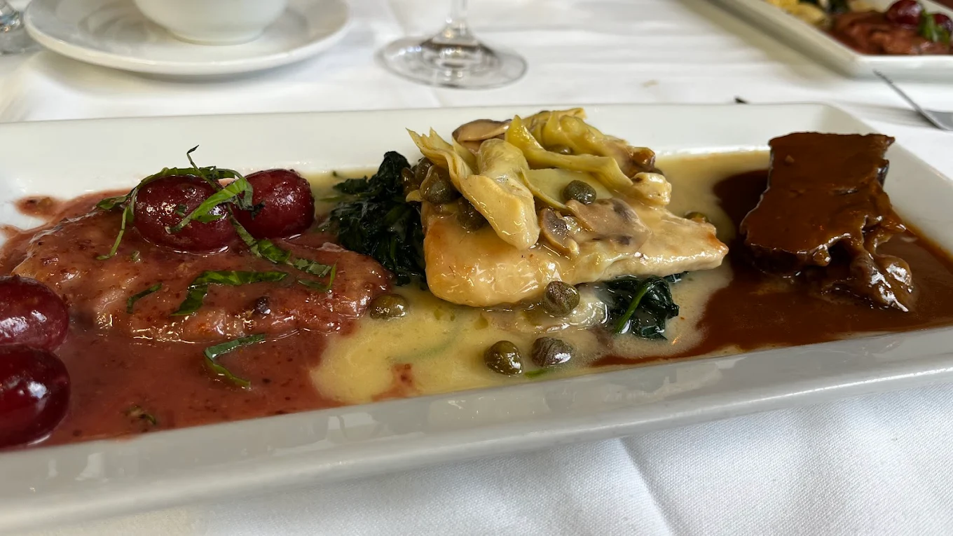 A rectangular white plate with three gourmet dishes: ravioli in red sauce with grapes, chicken in a creamy caper and artichoke sauce over spinach, and a piece of beef in brown gravy, set on a white tablecloth.