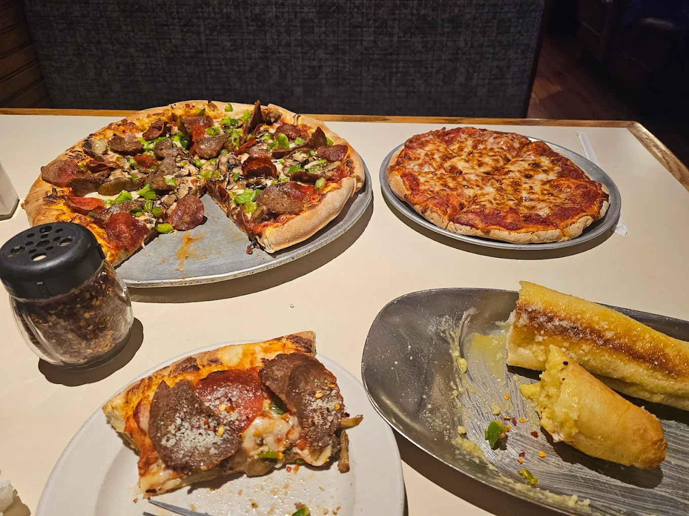 Two large pizzas sit on metal trays—one with pepperoni, beef, and green peppers; the other plain cheese. A shaker of pepper flakes and a plate with two breadsticks and a pizza slice are also on the table.