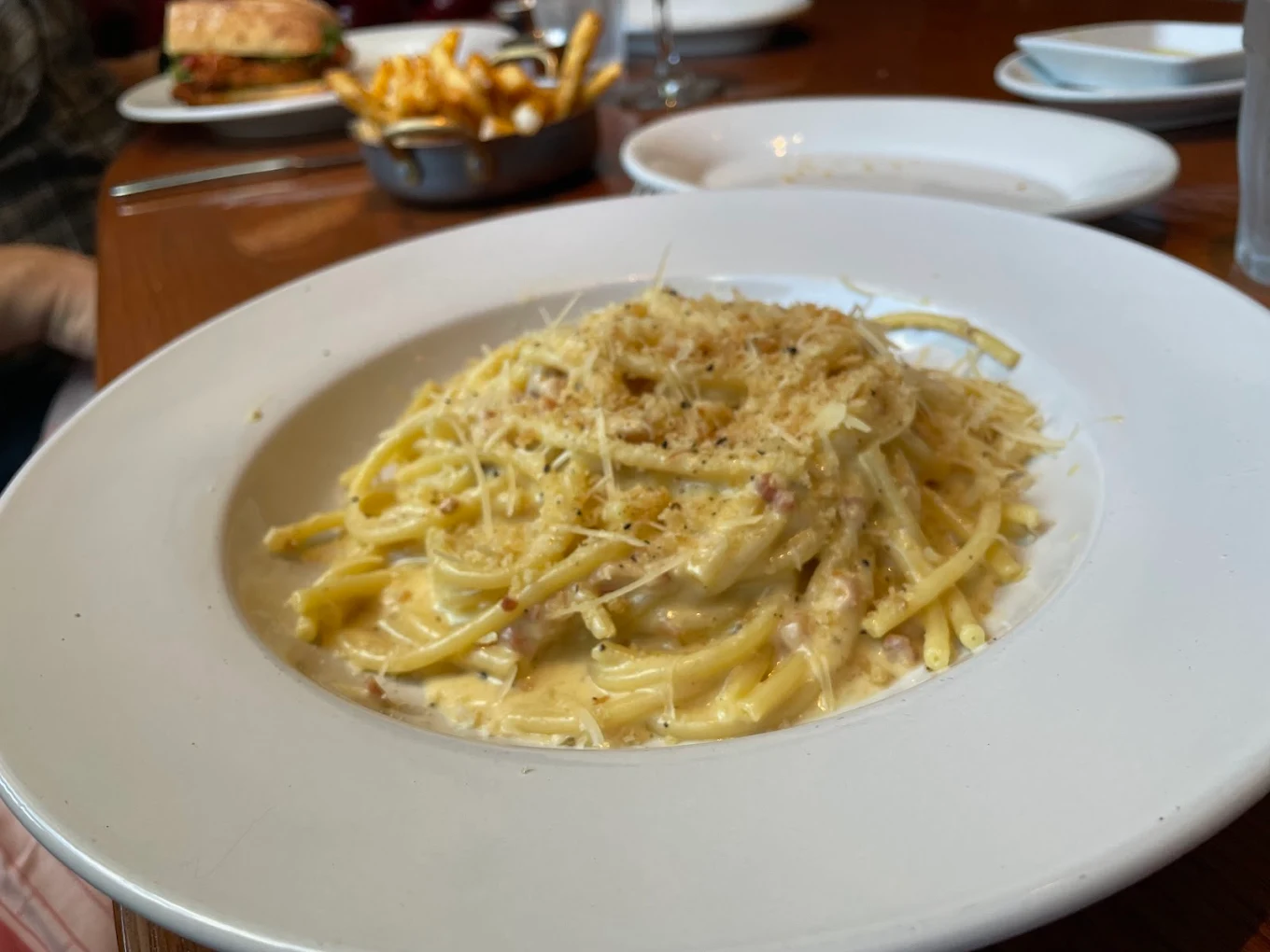 A white plate filled with creamy pasta topped with grated cheese sits on a wooden table, with fries, a sandwich, and empty dishes visible in the background.