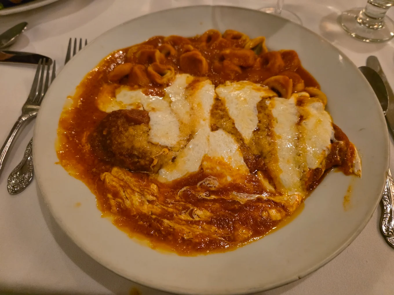 A plate of chicken parmesan topped with melted cheese and marinara sauce is served with a side of pasta in red sauce on a white tablecloth, surrounded by silverware.