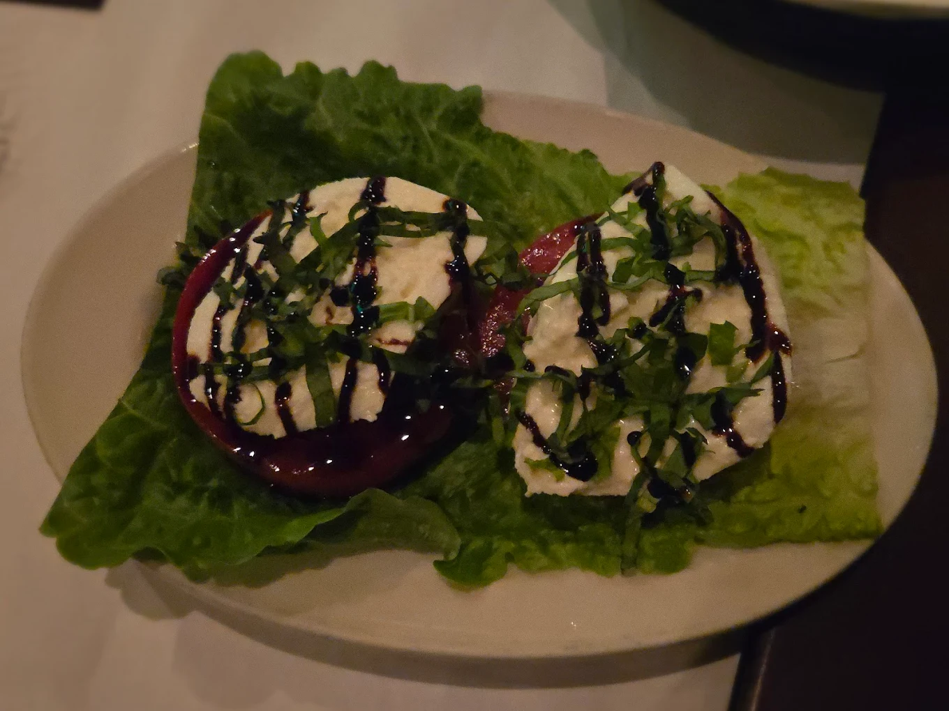 Two slices of tomato topped with mozzarella cheese, chopped basil, and a drizzle of balsamic glaze, served on a bed of leafy lettuce on an oval plate.