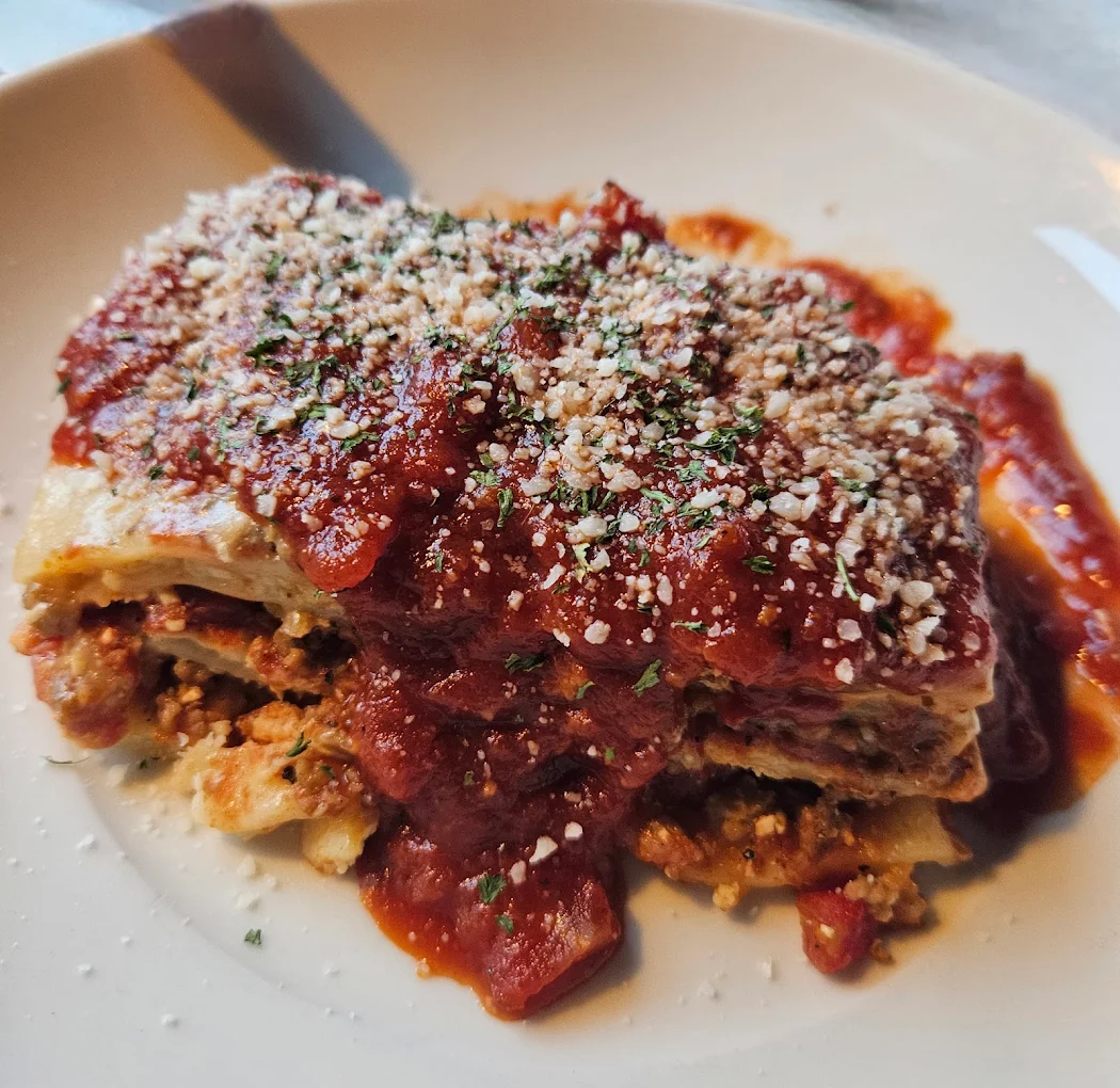 A slice of lasagna on a white plate, topped with tomato sauce, grated cheese, and sprinkled herbs. The layers of pasta, meat, and sauce are visible.