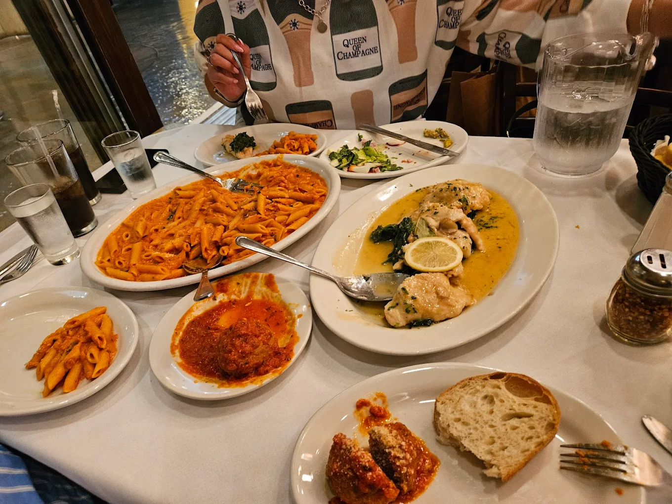 A table set for dinner with plates of penne pasta in red sauce, chicken in a lemon sauce with spinach, salad, bread, and glasses of water. A person in a patterned shirt is seated with a fork in hand.