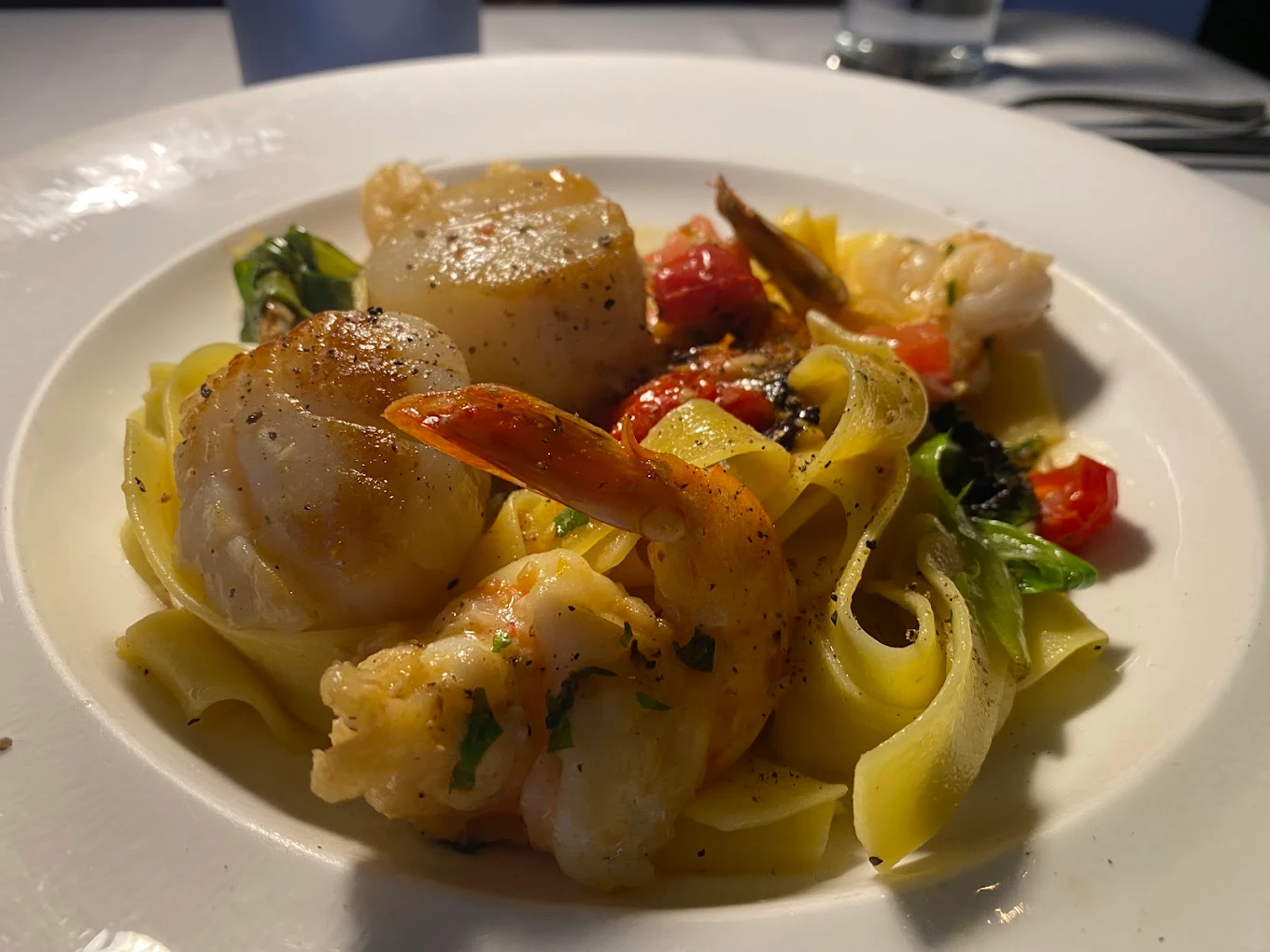 A close-up of a plate of pasta topped with seared scallops, shrimp, cherry tomatoes, and greens, served on a white dish with a glass of water and utensils in the background.