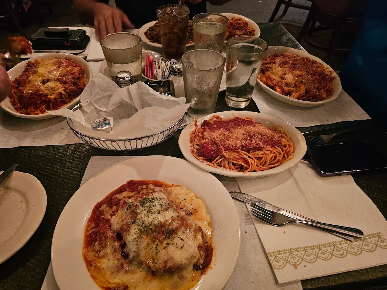 A restaurant table set with plates of pasta dishes topped with tomato sauce and cheese, glasses of water, a basket of bread with napkins, and utensils on napkins. Several people are seated around the table.