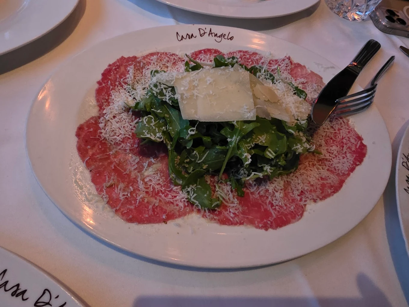 A plate of beef carpaccio topped with arugula, shaved Parmesan cheese, and grated cheese, served on a white plate with "Casa D'Angelo" written on the rim. A fork and knife are placed on the side.