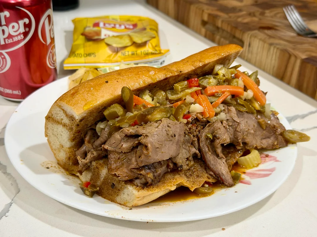 A loaded Italian beef sandwich with peppers on a plate, with Lay’s chips and a can of Dr Pepper in the background on a countertop.