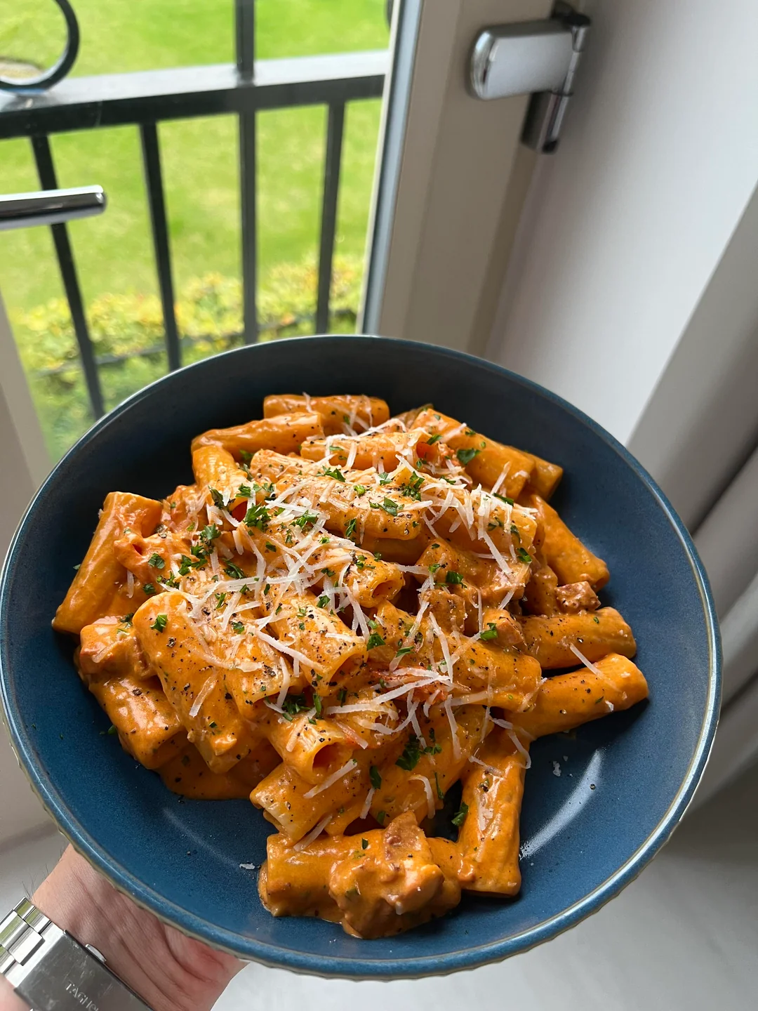 A blue bowl filled with creamy rigatoni pasta topped with grated cheese and herbs, held by a window with a view of a green lawn and black railing.