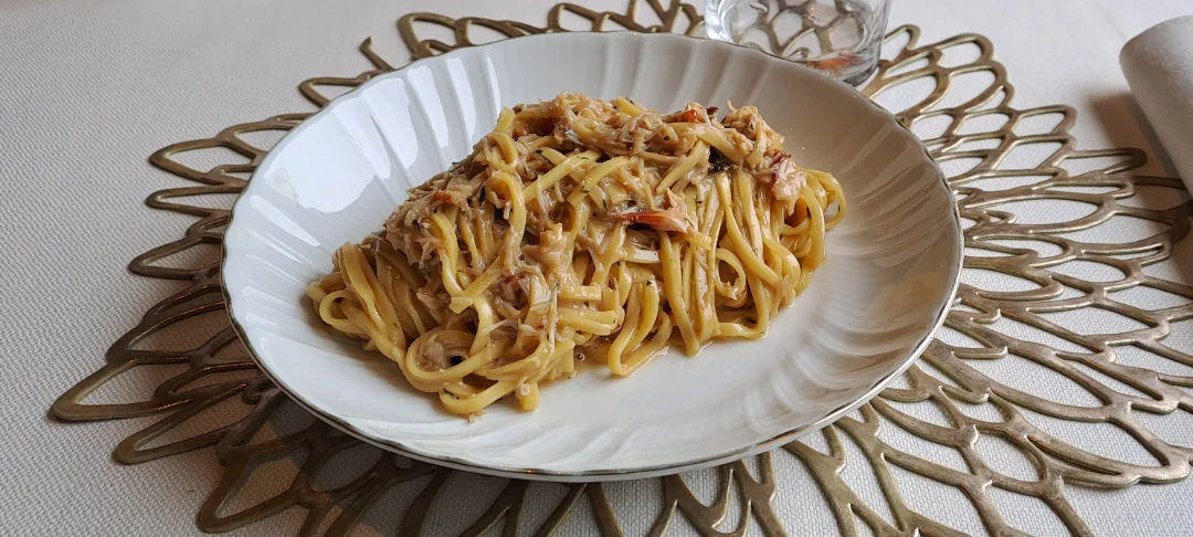 A high-angle close-up shot shows a plate of linguine with flaked crab meat in a light, creamy sauce, served in a fluted white bowl sitting on a gold, intricate, leaf-patterned placemat on a white tablecloth.