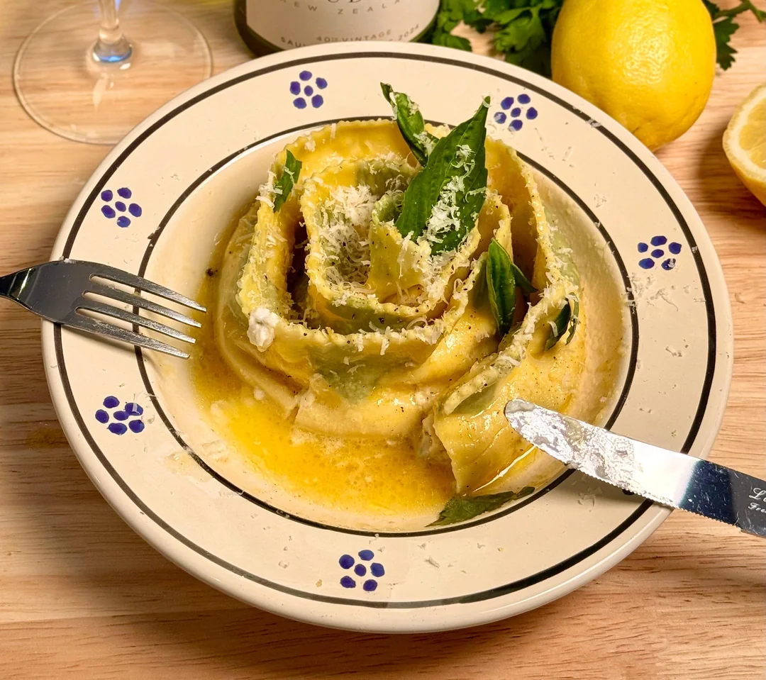 Homemade Italian food. A plate of rolled pasta topped with grated cheese and basil leaves sits in a creamy yellow sauce. A fork and knife rest on the plate. Lemon, herbs, and a wine glass are in the background.