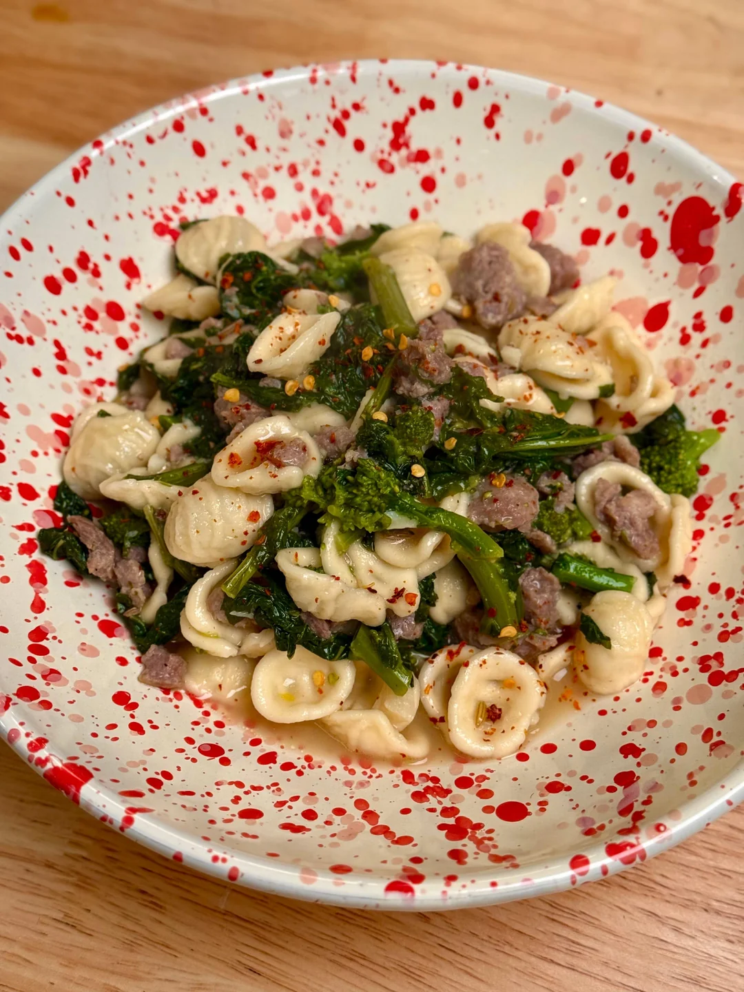 Orecchiette pasta with sausage, broccoli rabe, and red pepper flakes served in a white bowl with red splatter design, placed on a wooden table.