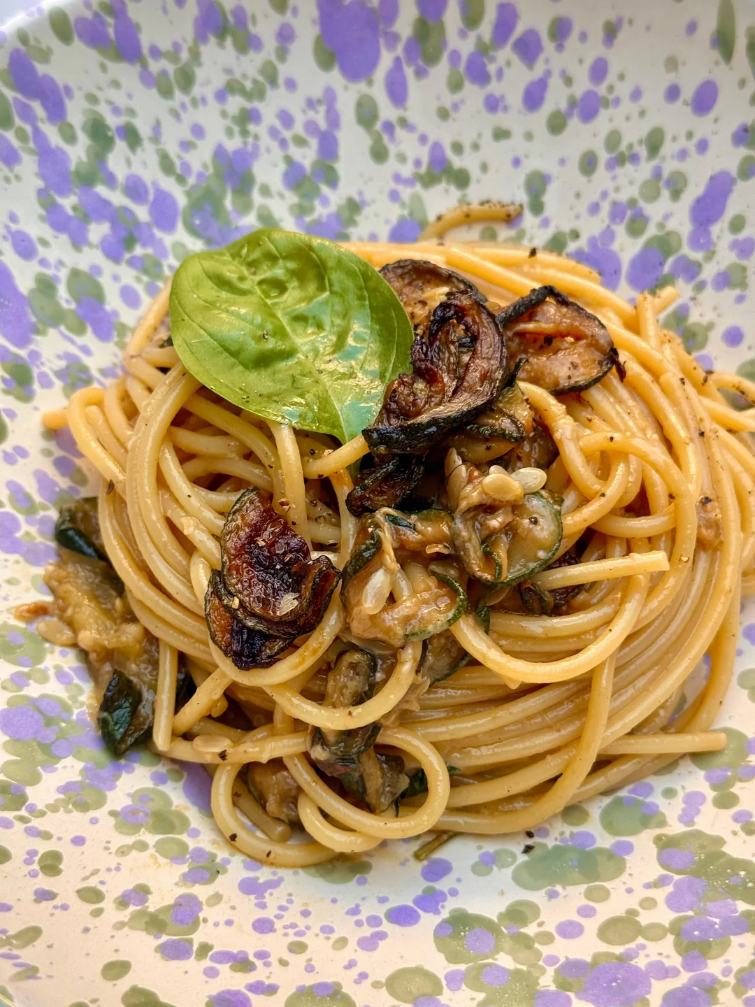 A plate of spaghetti with grilled zucchini slices, garnished with a fresh basil leaf, served on a speckled purple and green ceramic dish.