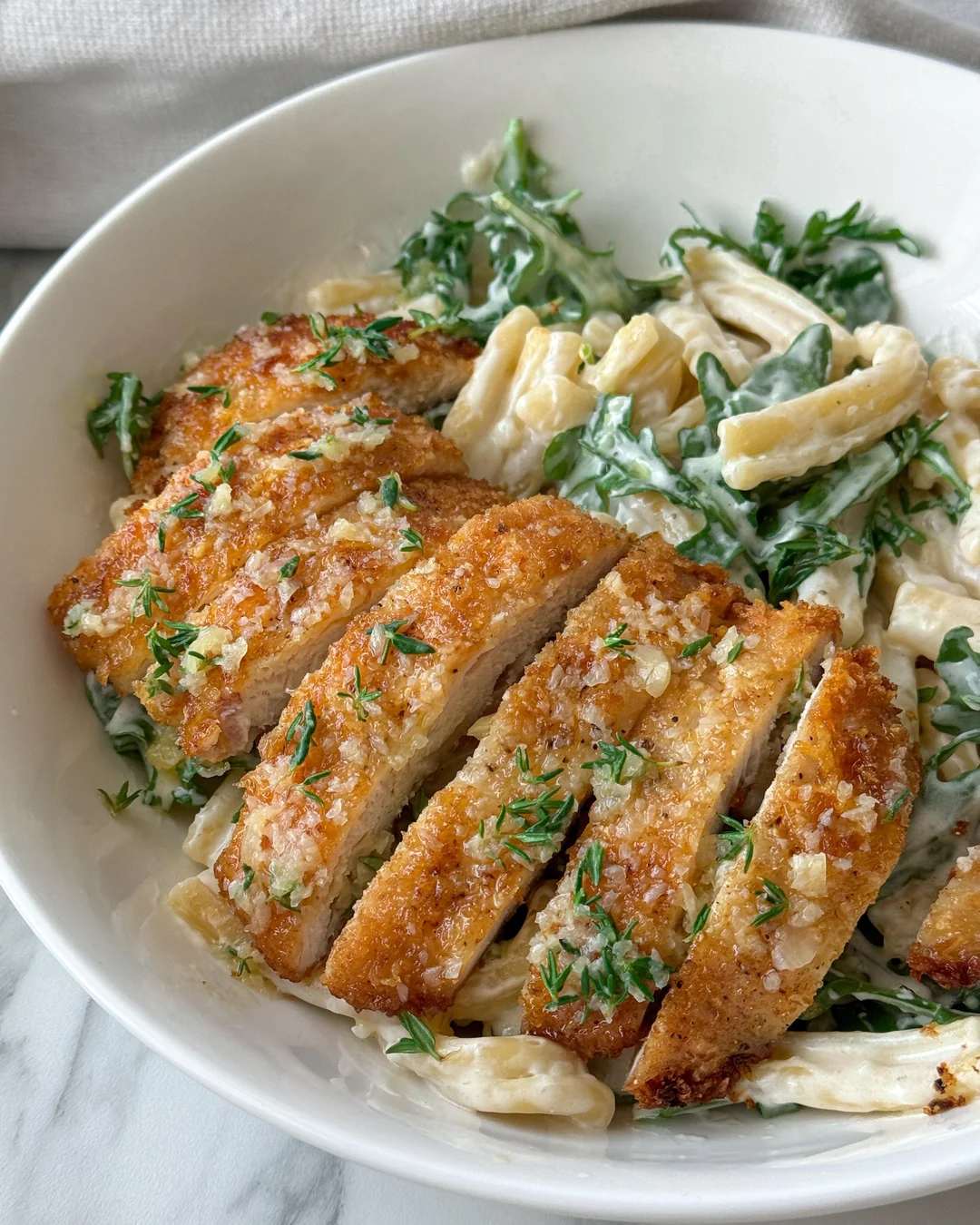 A white bowl with creamy pasta mixed with greens, topped with sliced, crispy breaded chicken and sprinkled with herbs.