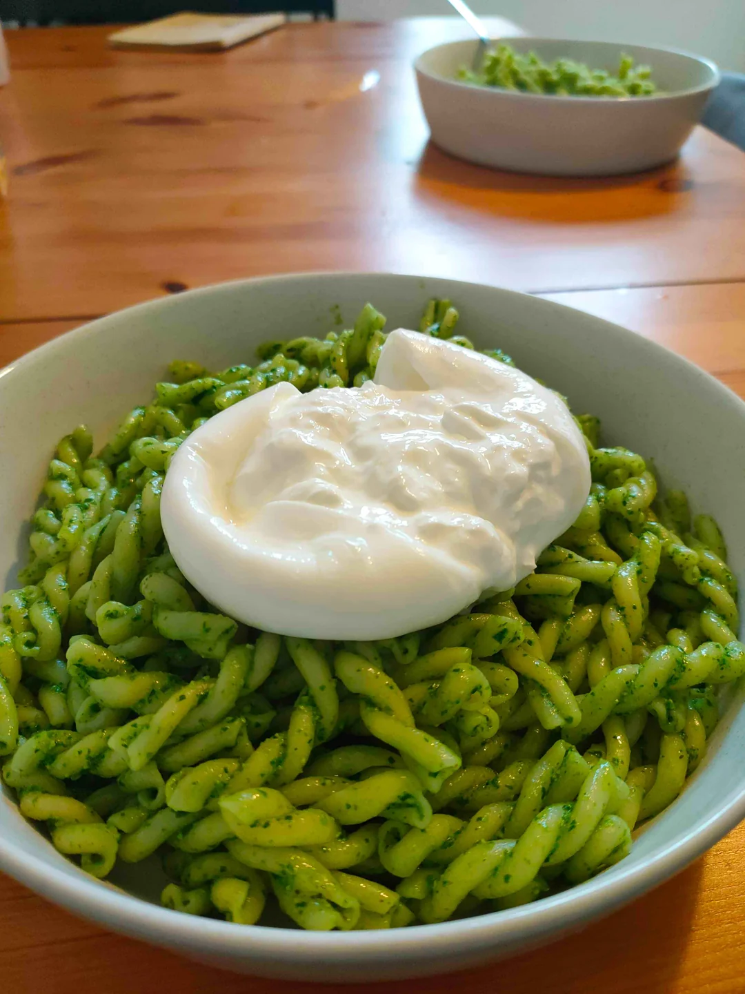 A close-up of a bowl of green pesto pasta topped with creamy burrata cheese, with another bowl of pasta blurred in the background on a wooden table.