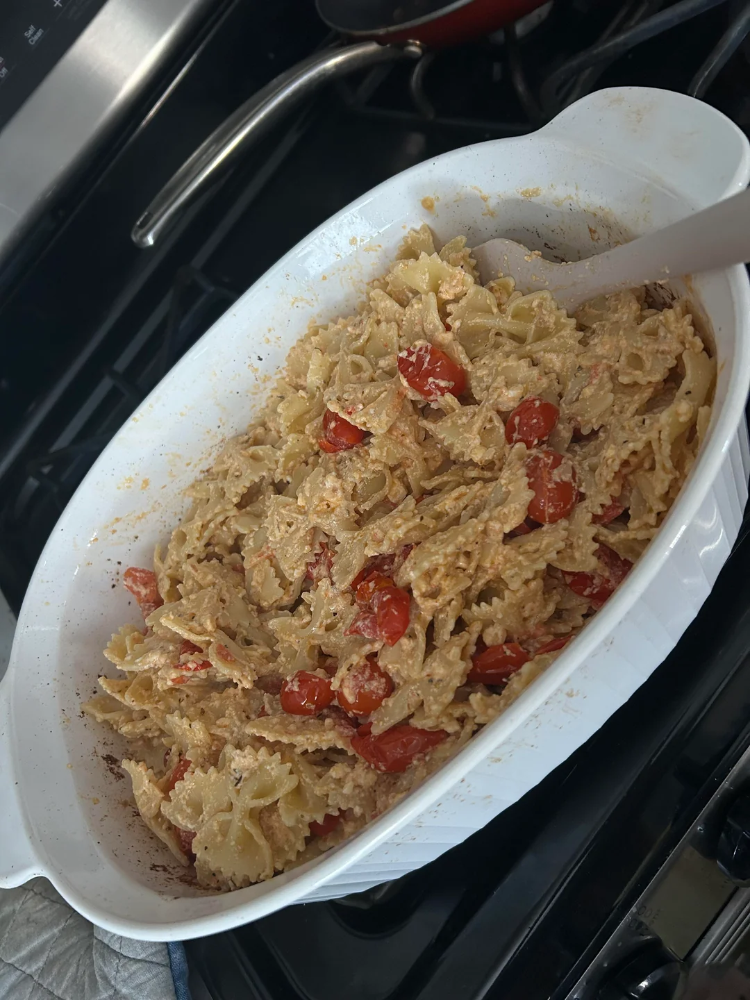 A white casserole dish filled with bowtie pasta, cherry tomatoes, and a creamy sauce, sitting on a stovetop with a spoon inside.