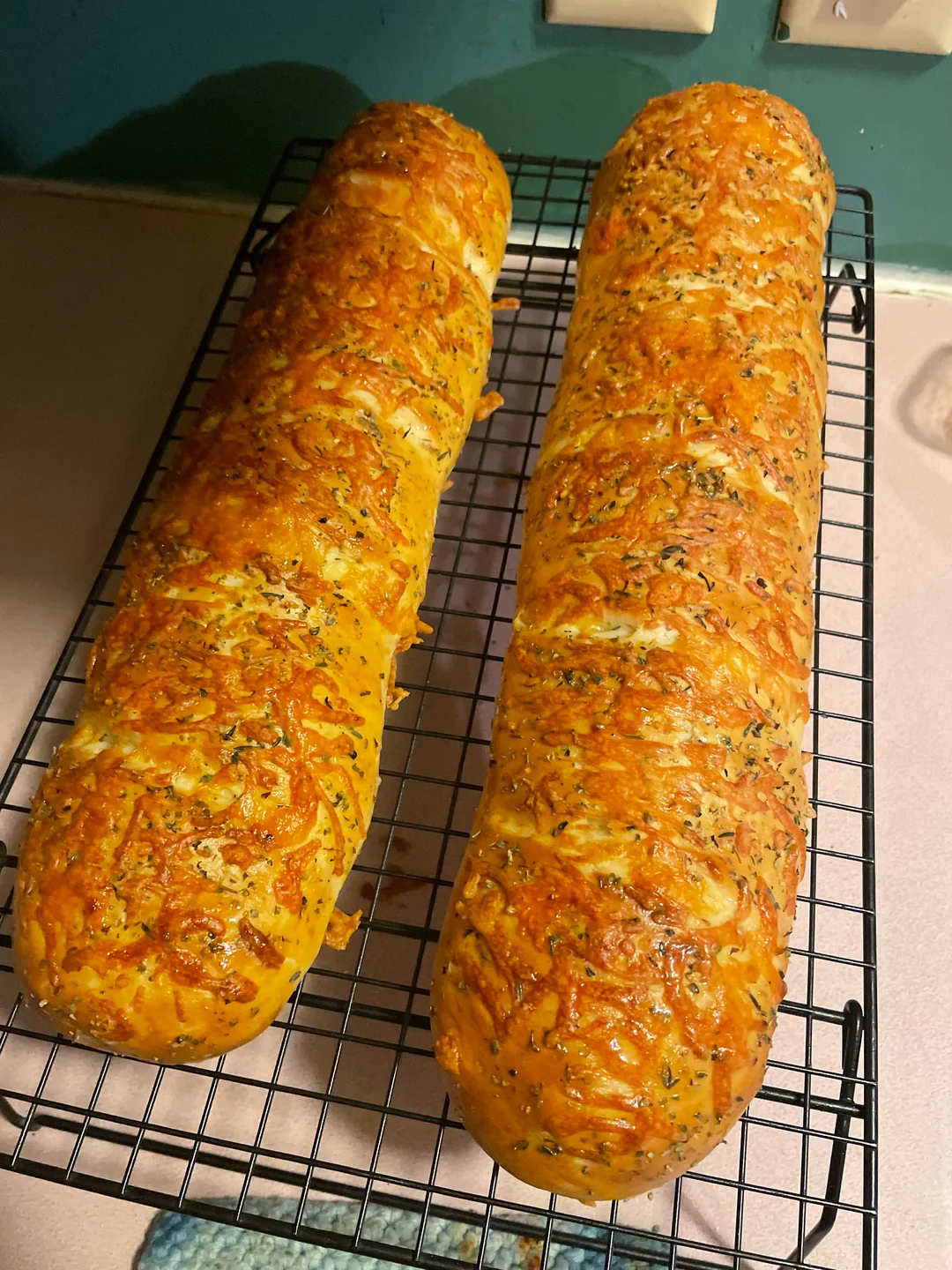 Two golden brown loaves of cheese and herb bread cool on a black wire rack, set on a kitchen counter with a teal wall and light-colored countertop in the background.