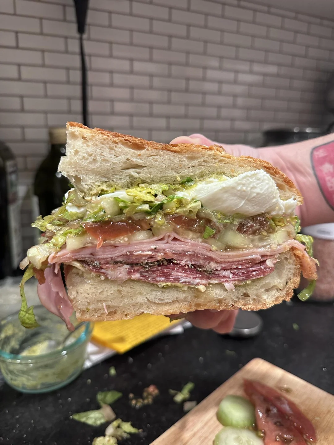 A hand holds a large Italian sub sandwich on crusty bread. The layers include thin-sliced deli meats, fresh mozzarella, chopped lettuce, tomato, and seasoned vinaigrette, with fresh cucumber and tomato slices visible on a cutting board below.