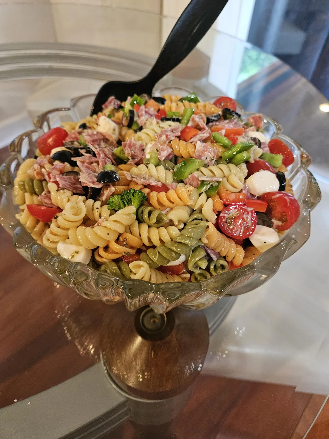 A glass bowl holds tri-color rotini pasta salad with cherry tomatoes, black olives, mozzarella, broccoli, salami, and green peppers. A black serving spoon rests in the bowl on a glass table.