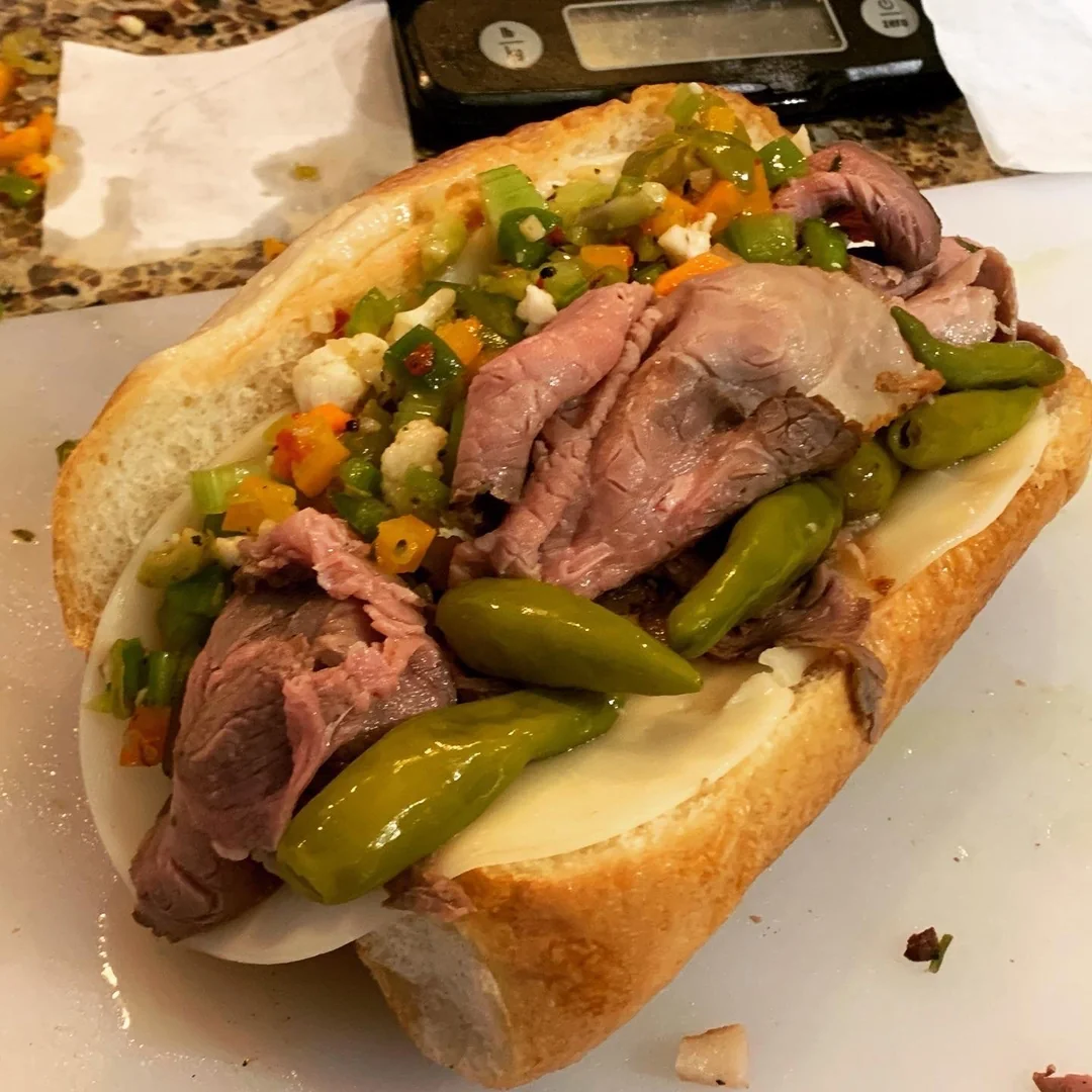 A sandwich on a hoagie roll filled with slices of roast beef, provolone cheese, green peppers, and a mix of chopped pickled vegetables. A digital scale is visible in the background.