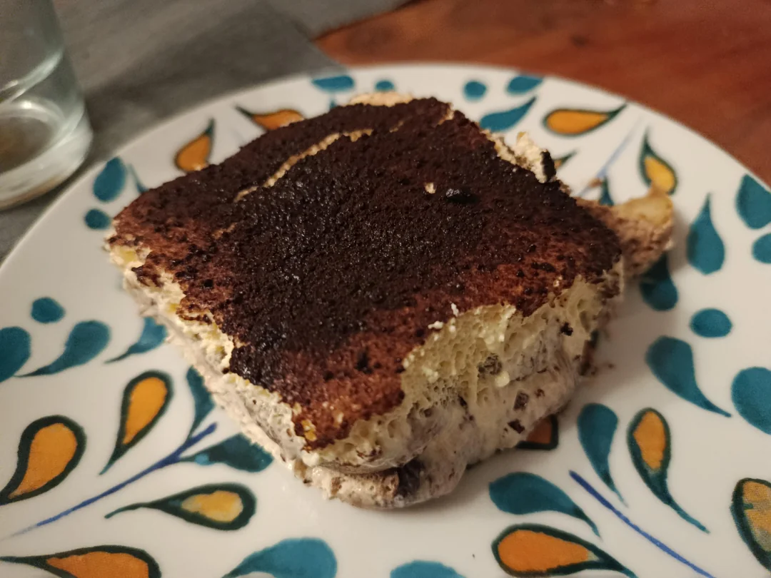A close-up of a slice of tiramisu on a decorative plate with a floral pattern, showing layers of cream and cocoa powder on top.